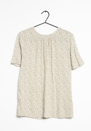 Blouse crème à manches courtes avec motif floral vert, encolure ronde, devant froncé, suspendue sur un cintre en métal noir.