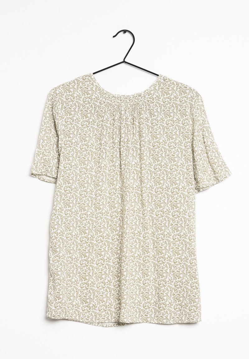 Blouse crème à manches courtes avec motif floral vert, encolure ronde, devant froncé, suspendue sur un cintre en métal noir.
