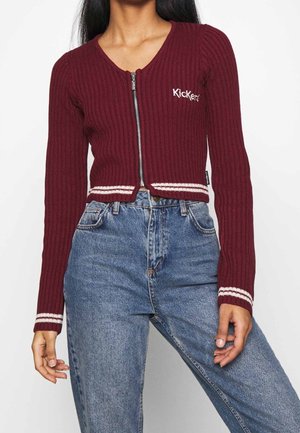 Femme portant un pull zippé côtelé bordeaux avec des rayures blanches aux poignets et à l'ourlet, associé à un jean en denim bleu taille haute.
