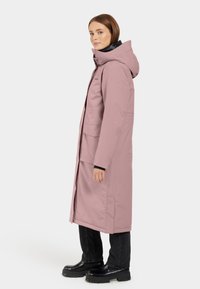 Parka lunga rosa con cappuccio, zip frontale, tasche laterali e interno nero. Indossata sopra pantaloni neri e stivali neri chunky.