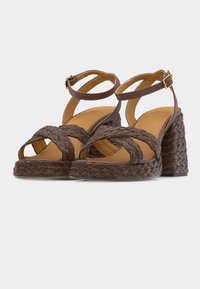 Καφέ βραχινα υποδήματα με χοντρό τακούνι, λουράκια στον αστράγαλο και χρυσά αγκράφια σε απλό φωτεινό φόντο.