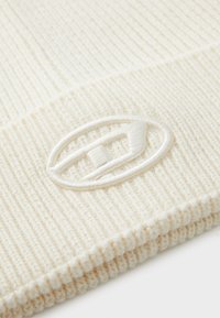 Bonnet en tricot couleur crème avec une texture côtelée et un revers replié. Présente un logo ovale brodé de la même couleur à l'avant.