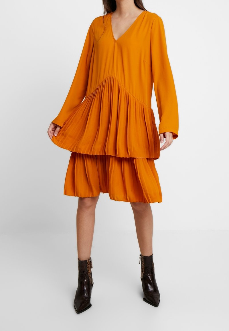 Femme portant une robe orange ample à manches longues avec des plis à volants, associée à des bottines marron foncé à motif peau de serpent.