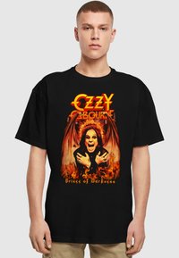Black cotton T-shirt featuring a colorful graphic of a figure with wings, flames, and "Ozzy Osbourne" text. The design emphasizes bold colors and imagery.