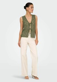 Zielony sweter bez rękawów z falbaniastego dołu, z zapięciem na przód. Noszony z kremowymi spodniami o szerokich nogawkach i białymi sandałami na obcasie.