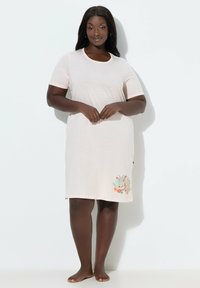 Mujer descalza, de pie, vistiendo un vestido corto de manga corta color rosa claro hasta la rodilla, con un pequeño diseño colorido de conejito y flores cerca del dobladillo.