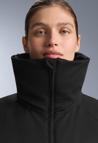 Crna jakna s visokim ovratnikom, glatkim vanjskim materijalom i prednjim patentnim zatvaračem. Ima teksturiranu unutarnju oblogu vidljivu na ovratniku.