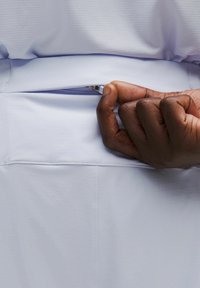Kéz, amely egy világos színű, rácsos mintázatú atlétikai rövidnadrág kis cipzáras zsebének nyílását nyitja.