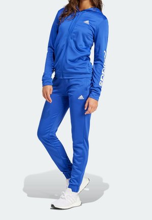 Kobieta ubrana w niebieski dres Adidas z kapturem i białe buty sportowe Adidas, stojąca z jedną nogą skrzyżowaną przed sobą na jasnym tle.