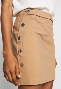 Tan mini-jurk met een getailleerde vorm, zijknoopsluiting met grote zilverkleurige knopen en een gladde stofstructuur.