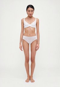 Conjunto de bralette y bikini de encaje blanco. El bralette tiene bordes festoneados, mientras que el bikini presenta un patrón floral. Textura suave y delicada.