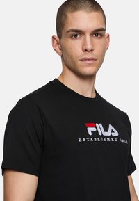 Camiseta de algodón negra con un prominente logo de FILA en rojo y blanco, con el texto "ESTABLECIDO EN 1911" bordado debajo. Mangas cortas, cuello redondo.