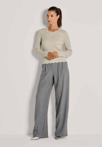 Frau, die vor einer hellen Wand steht und einen beigen Pullover mit gewelltem Saum, graue weite Hosen und silberne spitze Schuhe trägt.