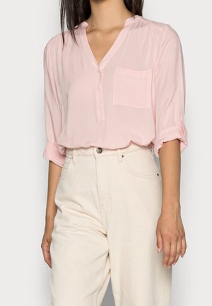 Frau trägt eine hellrosa Bluse mit Knöpfen und hochgekrempelten Ärmeln sowie eine cremeweiße, hochtaillierte Hose und steht vor einem einfarbigen Hintergrund.