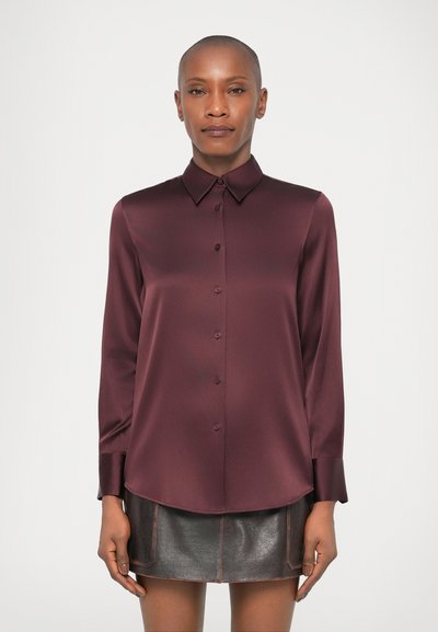 Femme portant une chemise satinée à manches longues de couleur bordeaux et une mini-jupe en cuir noire, se tenant devant un fond clair et uni.
