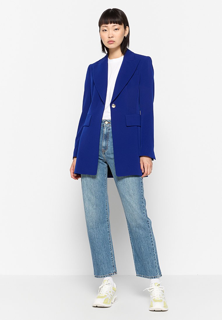 Royalblauer Blazer mit einem Knopf, Reverskragen und zwei Vorder Taschen, kombiniert mit hellblauen geraden Jeans und weißen Sneakers.