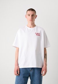 Balta medvilnė marškinėliai trumpomis rankovėmis, apvaliu kaklu ir raudonu "Calvin Klein" logotipu kairėje krūtinės pusėje. Dėvimi su mėlynais džinsais.