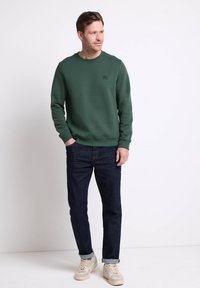 Felpa verde in tessuto testurizzato, con polsini a coste e collo rotondo; abbinata a jeans in denim scuro e sneakers chiare. Branding minimal visibile.