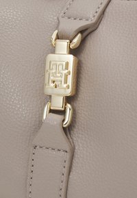 Detail popruhu světle šedé kožené kabelky s pozlaceným zapínáním a logem. Texturovaný povrch s viditelným prošíváním.