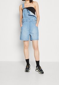 Salopette in denim azzurro chiaro con una spallina, dotata di tasca frontale e tasche laterali. Abbinata a una maglietta nera e scarpe da ginnastica.