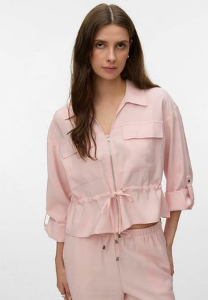 Młoda kobieta z długimi brązowymi włosami, ubrana w jasnoróżową kurtkę na zamek z wiązaniem w talii i pasujące spodnie, na jednolitym tle.