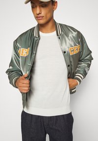 Green satin bomber jacket with orange embroidered lettering, striped ribbed cuffs, and a white crew neck shirt underneath. Plain dark trousers.