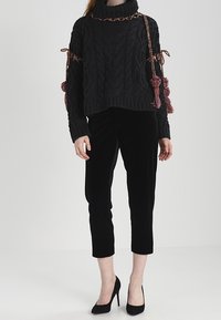 Svart kabelstickad tröja med hög krage och intrikata detaljer, kombinerad med svarta korta sammetsbyxor och svarta högklackade skor.