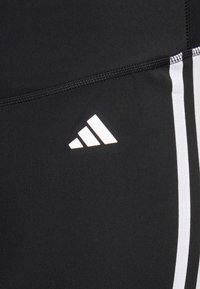 Czarne legginsy sportowe z białym logo Adidas, z białymi paskami biegnącymi wzdłuż boków i gładkim, elastycznym materiałem.