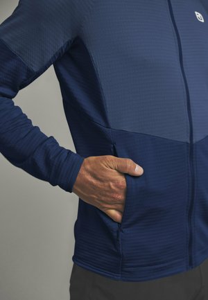 Mann trägt marineblaue Zip-Up-Jacke mit strukturiertem Stoff, Hand steckt in seitlicher Reißverschlusstasche, vor neutralem Hintergrund.