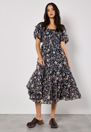 Navy dress with floral pattern, puff sleeves, and smocked bodice. Features a tiered skirt and a relaxed fit. Worn with dark clogs.