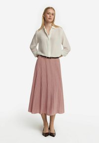 Vit skjorta med lång ärm, kombinerad med en plisserad rosa kjol. Kjolen har en glänsande yta och ett solid svart bälte. Bruna spetsiga skor.