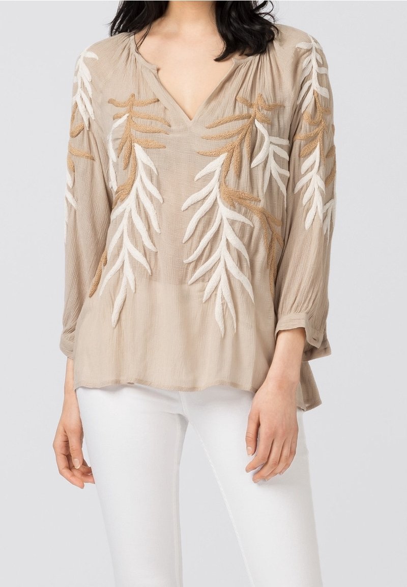 Femme portant un chemisier beige à manches longues avec une broderie de feuilles blanches et marron, assorti à un pantalon blanc, debout devant un fond uni.