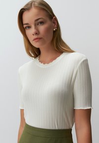 Biały prążkowany top dzianinowy z falbaniastego dekoltem i krótkimi rękawami, zestawiony z zieloną spódnicą. Tkanina ma miękką teksturę.