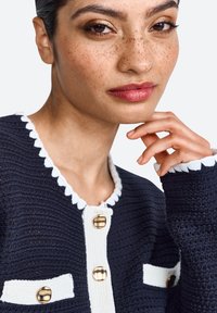 Cardigan lavorato a maglia di colore blu navy con bordi bianchi, dotato di bottoni dorati e tasche a toppa, texture strutturata e dettagli del colletto a scallop.