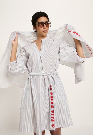 Person trägt ein gestreiftes, mit Gürtel versehenes Kleid und hält einen passenden gestreiften Schal mit rotem Text, dazu große brauntönte Sonnenbrillen als Accessoire.