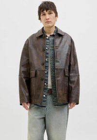 Brown leather jacket with a collared neckline, button closure, and two front pockets. Underneath, a blue denim shirt and grey jeans.