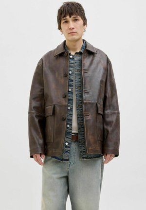 Brown leather jacket with a collared neckline, button closure, and two front pockets. Underneath, a blue denim shirt and grey jeans.