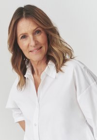Camicia bianca con bottoni, dal tessuto morbido, con maniche arrotolate e colletto strutturato, indossata da una donna con capelli mossi fino alle spalle.