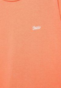 Gros plan d’un tissu couleur corail avec un petit logo brodé blanc « Swtz » près du centre supérieur.