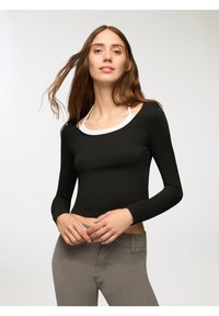 Crop top nero a maniche lunghe con colletto bianco, realizzato in tessuto liscio. La modella lo indossa con pantaloni grigi testurizzati.