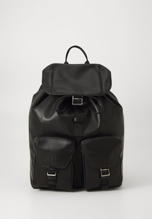 Sac à dos en cuir noir avec rabat supérieur, boucle à l'avant, fermeture à cordon, et deux poches avant avec boucles sur fond blanc.