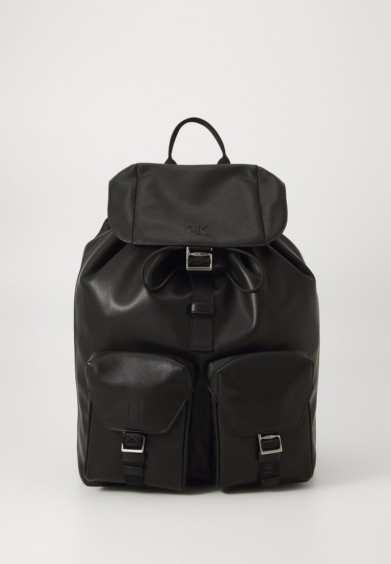 Sac à dos en cuir noir avec rabat supérieur, boucle à l'avant, fermeture à cordon, et deux poches avant avec boucles sur fond blanc.