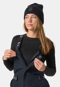 Czarna czapka beanie i dopasowany czarny top z długim rękawem pod granatowymi ogrodniczkami z regulowanymi ramiączkami i detalami w postaci zamków. Widoczny tekst na ramiączkach.