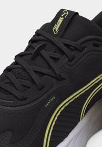 Μαύρο παπούτσι τρεξίματος Puma με μαύρα κορδόνια, κίτρινα στοιχεία και λευκή σόλα, με ετικέτα «PWRTAPE» στην πλαϊνή επιφάνεια.