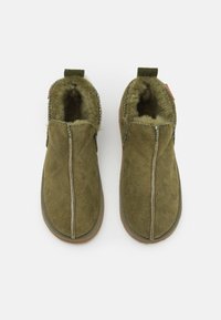 Para oliwkowych zamszowych kapci z miękkim, puszystym wykończeniem i gumowymi podeszwami, widziana z góry na białym tle.