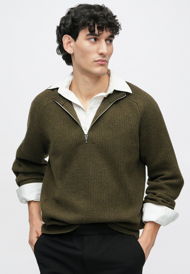 Groene geribbelde trui met een halve rits halslijn, lange mouwen en een witte blouse eronder, gedragen met zwarte broek.