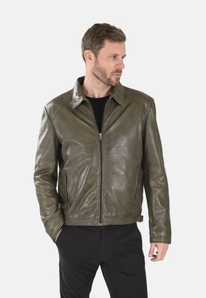 Homme barbu portant une veste en cuir vert olive et un pantalon noir, debout avec une main dans la poche, regardant vers sa droite.