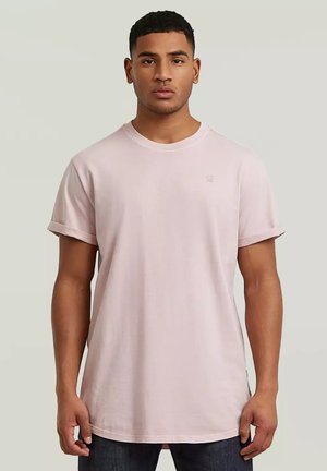 Giovane uomo che indossa una t-shirt a maniche corte rosa chiaro tinta unita e pantaloni scuri, in piedi davanti a uno sfondo neutro.