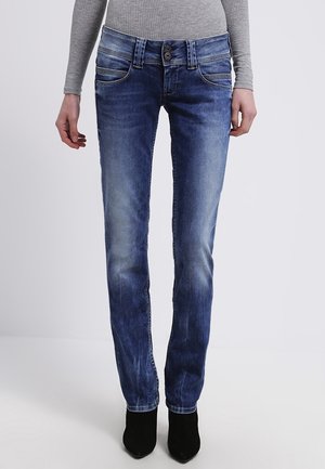 Personne portant un jean slim bleu délavé et des bottines noires, associées à un haut gris à manches longues côtelé, debout devant un fond uni.