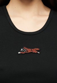 Κοντινό πλάνο μαύρου ριπ μπλουζιού με κέντημα από χάντρες, κόκκινο και λευκό, σε σχήμα σκύλου κοντά στη λαιμόκοψη, στο πάνω μέρος του στήθους ενός ατόμου.